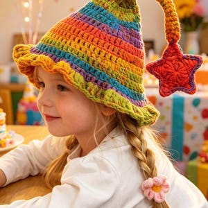May include: A crocheted hat featuring a vibrant rainbow-striped pattern with a wide, ruffled brim. The hat has a pointed top that curves downward, ending in a dangling, textured red star. The hat is worn by a person with blonde hair in braids.