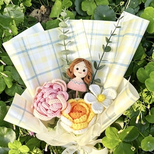 May include: A crochet bouquet with a doll, pink and yellow flowers, and a white flower. The bouquet is wrapped in yellow plaid paper and tied with a white ribbon. The arrangement is surrounded by green foliage.