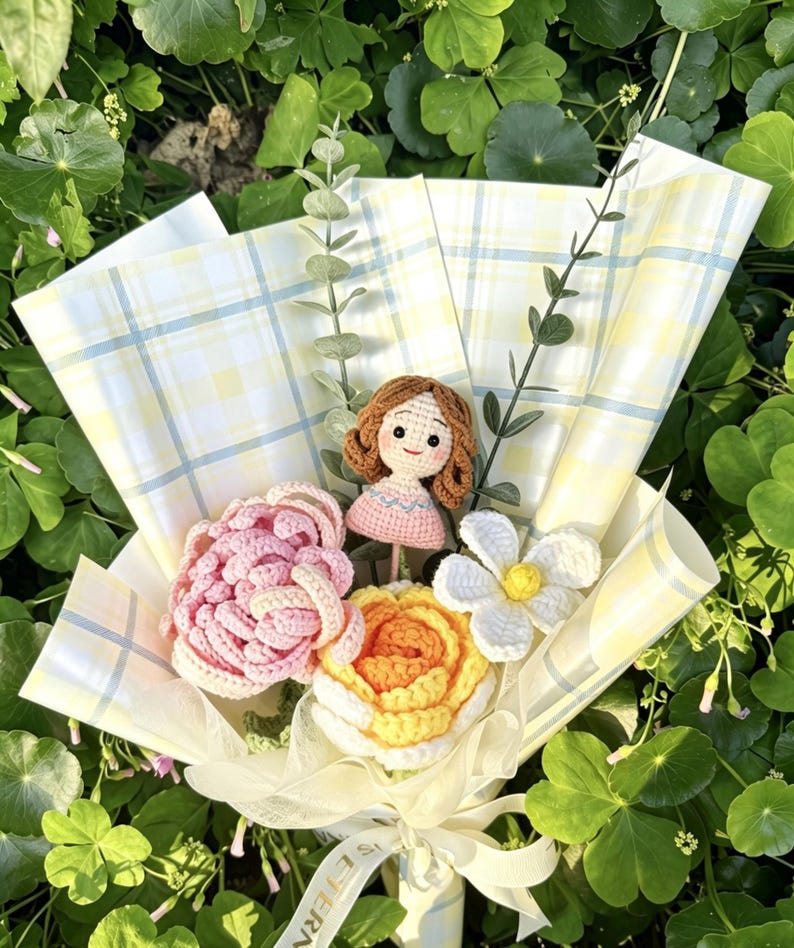 May include: A crochet bouquet with a doll, pink and yellow flowers, and a white flower. The bouquet is wrapped in yellow plaid paper and tied with a white ribbon. The arrangement is surrounded by green foliage.