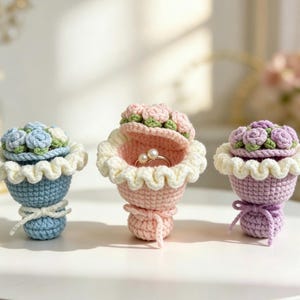 May include: Three handmade crochet ring boxes shaped like bouquets. One is blue with blue and white flowers, one is pink with pink roses and a ring inside, and the third is purple with purple roses.