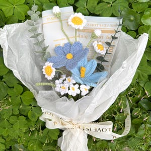 May include: A handmade crochet flower bouquet in blue, white, and yellow, wrapped in white paper. The bouquet features daisies and other flower types, tied with a ribbon that reads "LOVE IS". The background is green foliage.