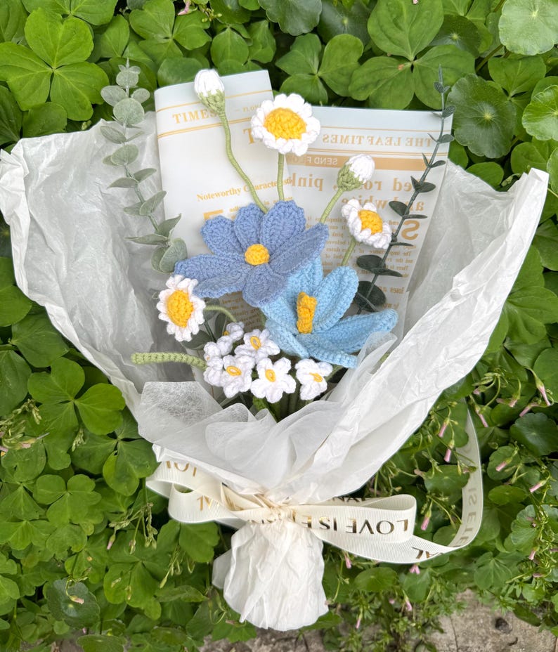May include: A handmade crochet flower bouquet in blue, white, and yellow, wrapped in white paper. The bouquet features daisies and other flower types, tied with a ribbon that reads "LOVE IS". The background is green foliage.