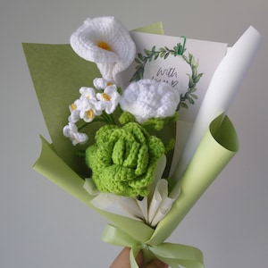 May include: A handmade crochet bouquet featuring white calla lilies, small white flowers, and a green succulent. The bouquet is wrapped in green and white paper, tied with a green ribbon, and includes a card that says "With love".
