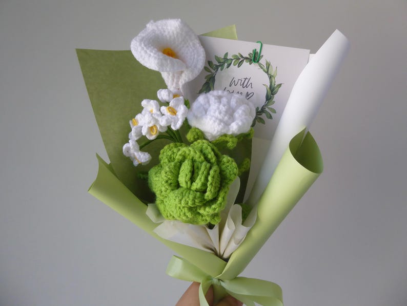 May include: A handmade crochet bouquet featuring white calla lilies, small white flowers, and a green succulent. The bouquet is wrapped in green and white paper, tied with a green ribbon, and includes a card that says "With love".