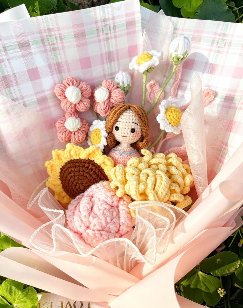 May include: A handcrafted crochet bouquet with pink, yellow, and white flowers, including daisies, sunflowers, and a large pink rose. A small doll is nestled among the flowers, all wrapped in pink and plaid paper.