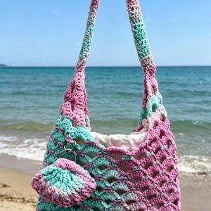 May include: A crochet shoulder bag featuring a scalloped pattern in variegated pink and mint green yarn. The bag includes a single strap, a solid white fabric interior lining, and a small matching circular crochet charm attached by a metal ring.