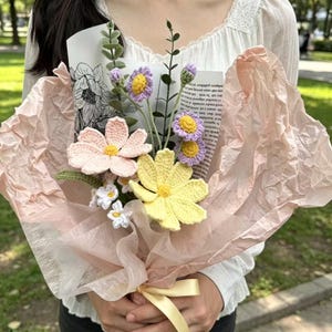 May include: A handcrafted bouquet of crochet flowers in pastel shades of pink, yellow, and purple, presented in pink tissue paper and tied with a yellow ribbon. The bouquet features different flower designs and green stems, offering a unique floral display.