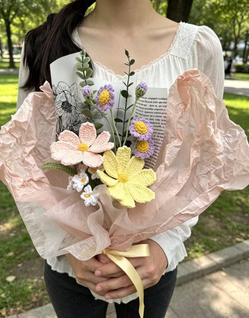 May include: A handcrafted bouquet of crochet flowers in pastel shades of pink, yellow, and purple, presented in pink tissue paper and tied with a yellow ribbon. The bouquet features different flower designs and green stems, offering a unique floral display.