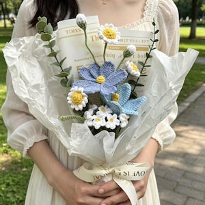 May include: A handmade crochet flower bouquet featuring blue and white daisies, wrapped in white paper. The bouquet is tied with a cream ribbon that reads "LOVE IS ETERNAL". The bouquet includes greenery and a newspaper clipping.