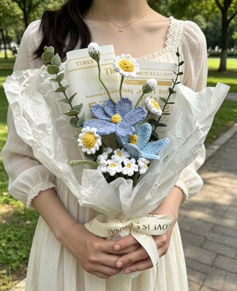 May include: A handmade crochet flower bouquet featuring blue and white daisies, wrapped in white paper. The bouquet is tied with a cream ribbon that reads "LOVE IS ETERNAL". The bouquet includes greenery and a newspaper clipping.