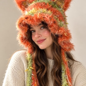 May include: A fuzzy, textured hat featuring two pointed cat-like ears on top. The hat is crafted from yarn in shades of bright orange, lime green, and yellow, with long, dangling braided ties extending from the sides of the head covering.