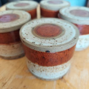 May include: A collection of handmade ceramic jars with lids. The jars feature a speckled off-white top, a band of reddish-brown, and a speckled off-white base. The lids have a reddish-brown center. The jars are arranged on a light brown wooden surface.