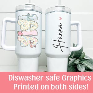 Puede incluir: Dos vasos aislados blancos con asas. Uno presenta el nombre "Hanna" en cursiva con un peque&ntilde;o coraz&oacute;n rosa. El otro tiene la palabra "DAD" en un dise&ntilde;o floral y pastel. Ambos tienen una tapa transparente y una pajita.