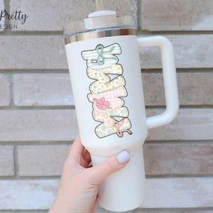Puede incluir: Vaso t&eacute;rmico blanco con asa, con la palabra "MOM" en un dise&ntilde;o apilado. Cada letra est&aacute; decorada con un patr&oacute;n floral y un lazo diferentes. El vaso tiene una tapa transparente y una pajita.