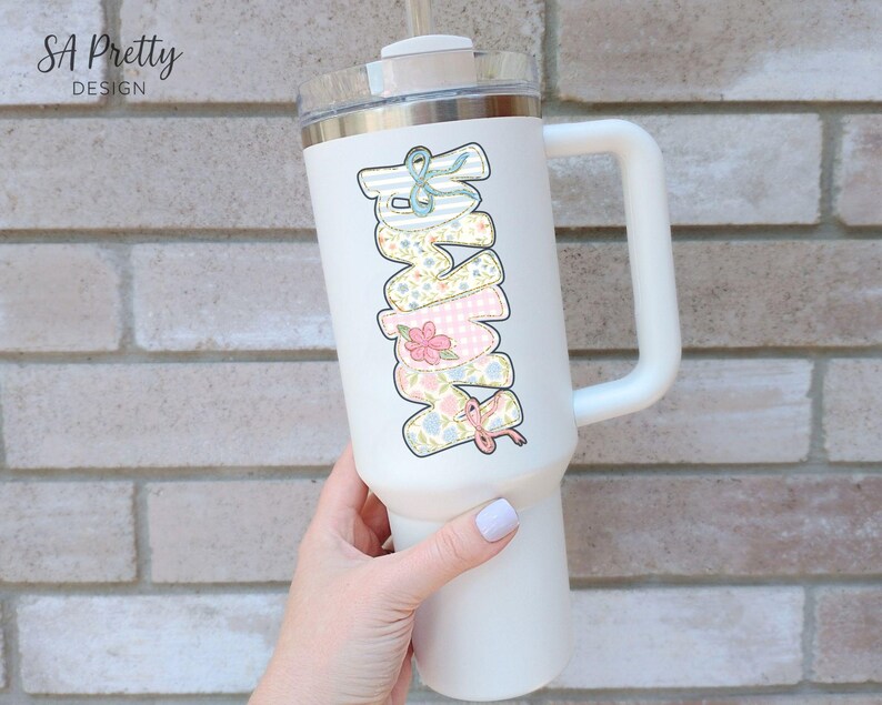 Puede incluir: Vaso t&eacute;rmico blanco con asa, con la palabra "MOM" en un dise&ntilde;o apilado. Cada letra est&aacute; decorada con un patr&oacute;n floral y un lazo diferentes. El vaso tiene una tapa transparente y una pajita.
