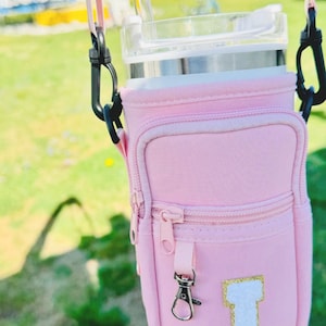 Puede incluir: Bolsa aislante rosa para vaso con cremallera y un bolsillo peque&ntilde;o. La bolsa tiene una letra "J" blanca con purpurina dorada en la parte delantera. La bolsa tiene una correa negra con un clip plateado.