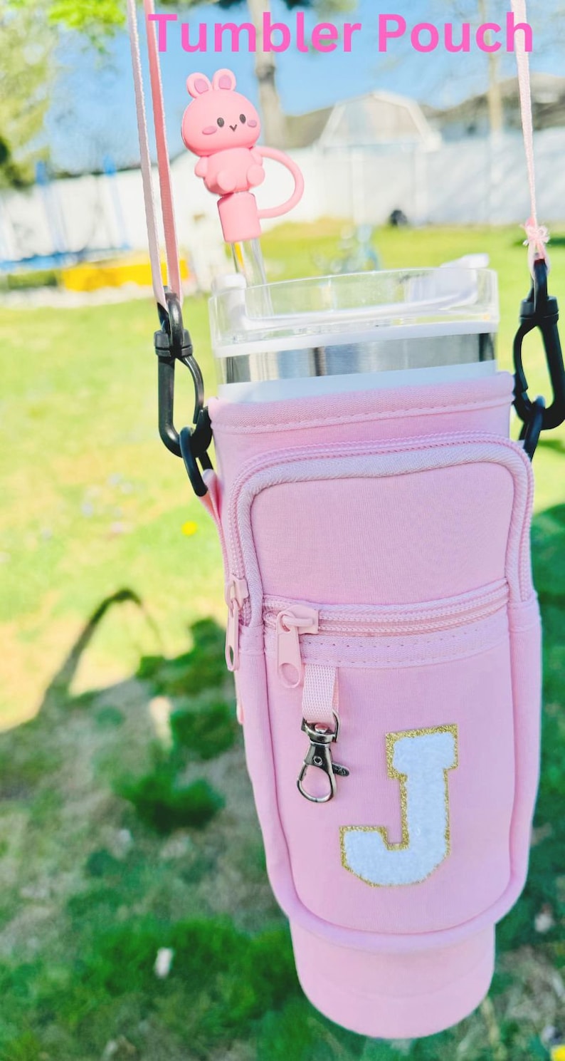 Puede incluir: Bolsa aislante rosa para vaso con cremallera y un bolsillo peque&ntilde;o. La bolsa tiene una letra "J" blanca con purpurina dorada en la parte delantera. La bolsa tiene una correa negra con un clip plateado.