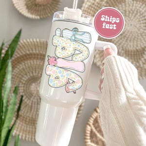 Puede incluir: Vaso blanco con asa, con un dise&ntilde;o floral y de lazo que deletrea "Love" en colores pastel. El vaso tiene una tapa transparente y una pajita. Un c&iacute;rculo rosa con las palabras "Ships fast" es visible.