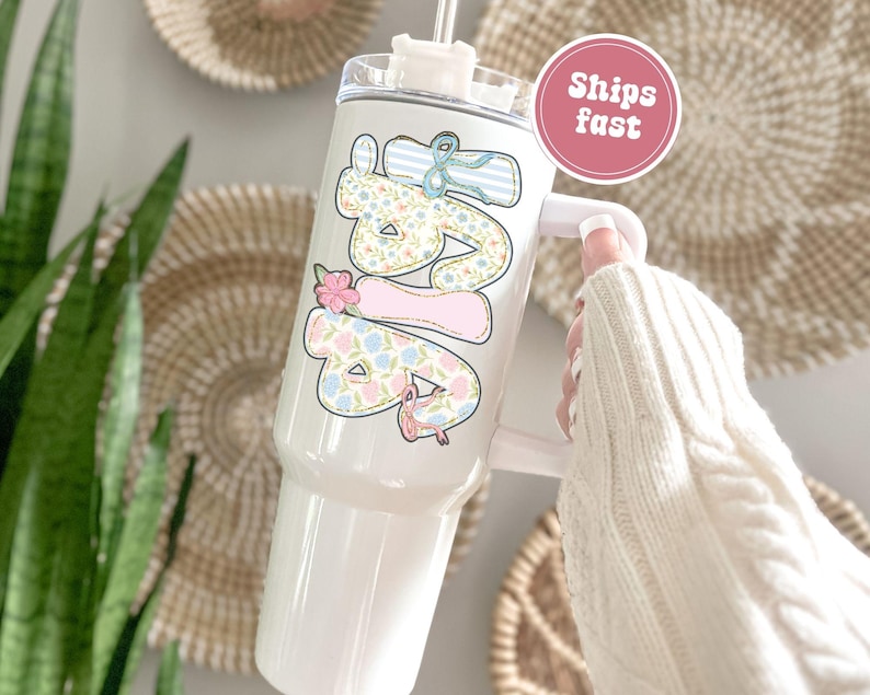 Puede incluir: Vaso blanco con asa, con un dise&ntilde;o floral y de lazo que deletrea "Love" en colores pastel. El vaso tiene una tapa transparente y una pajita. Un c&iacute;rculo rosa con las palabras "Ships fast" es visible.
