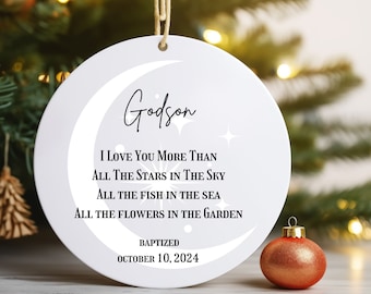 May include: A white ceramic Christmas ornament with a crescent moon design and the word "Godson" in script. The ornament includes the text "I Love You More Than..." and "Baptized October 10, 2024." A gold-colored ornament sits to the right.