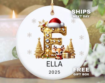 Puede incluir: Adorno navideño blanco con una letra "E" dorada adornada con pedrería, un gorro de Papá Noel y un reno. El adorno incluye el nombre "ELLA" y el año "2025". Copos de nieve y árboles de Navidad completan el diseño.