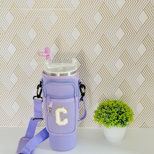 Puede incluir: Un vaso t&eacute;rmico morado con un bolsillo con cremallera y una correa para el hombro. El vaso tiene una letra "C" blanca en la parte delantera.