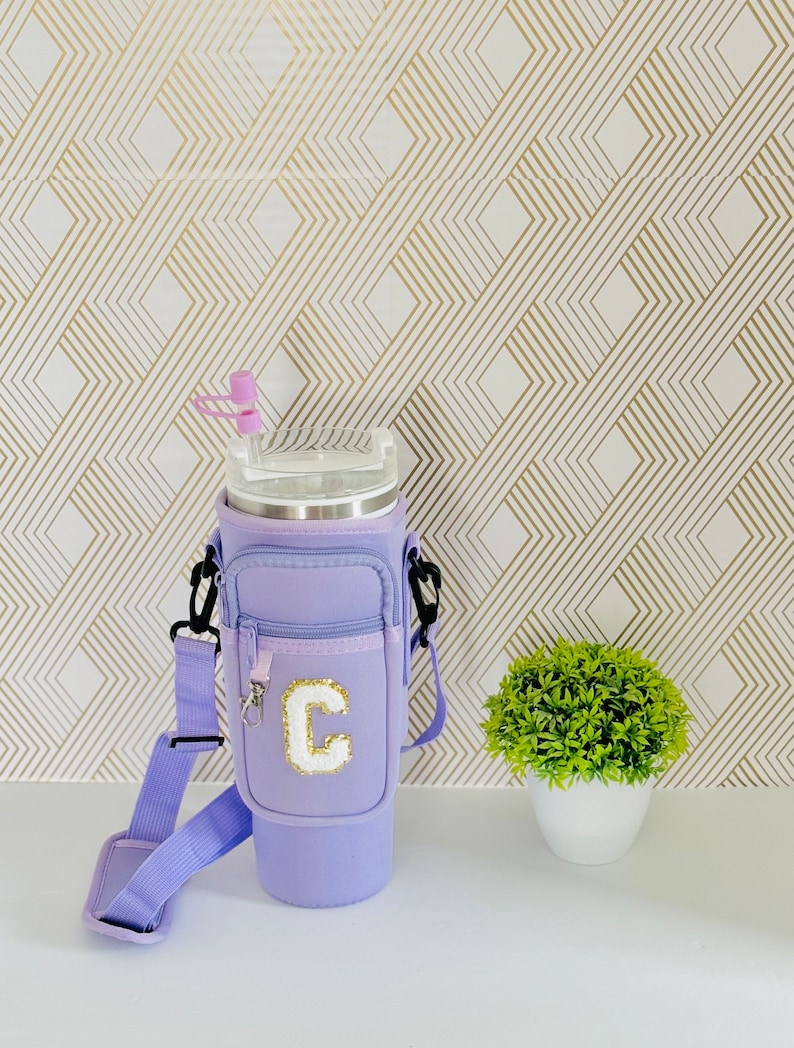 Puede incluir: Un vaso t&eacute;rmico morado con un bolsillo con cremallera y una correa para el hombro. El vaso tiene una letra "C" blanca en la parte delantera.