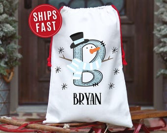 Puede incluir: Bolsa de regalo blanca con cordón y diseño de muñeco de nieve con la letra "B" y el nombre "Bryan". El muñeco de nieve tiene un sombrero de copa negro, bufanda azul y nariz naranja. Copos de nieve alrededor del diseño. Pegatina roja "SHIPS FAST".