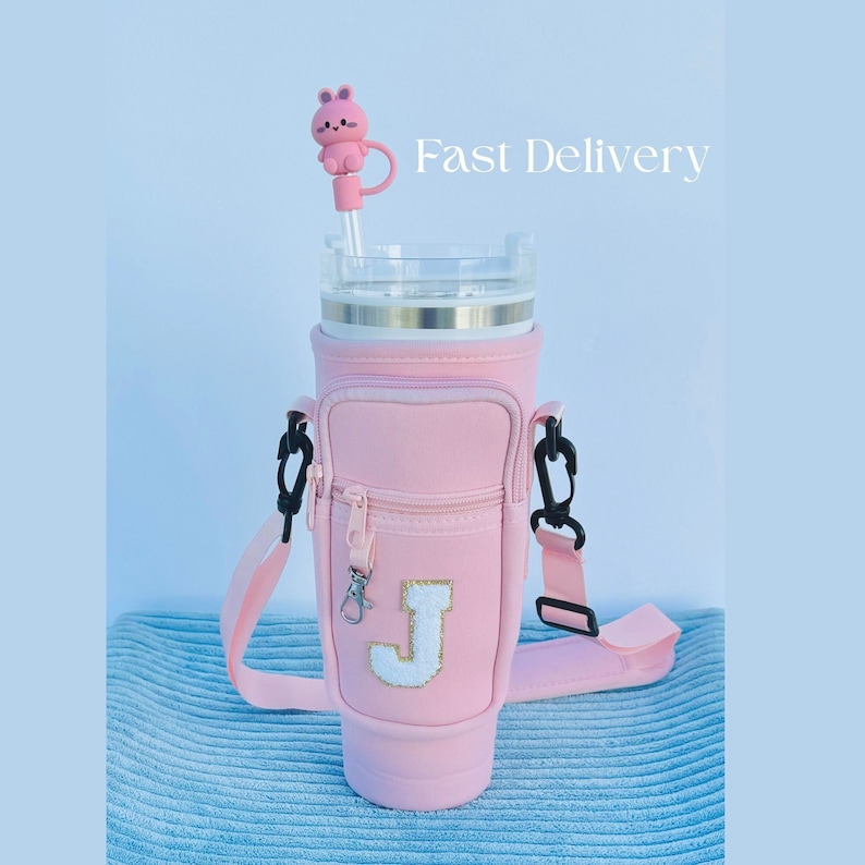 Puede incluir: Un vaso t&eacute;rmico rosa con un bolsillo con cremallera y una correa. El vaso tiene una letra "J" blanca en la parte delantera y un tap&oacute;n de pajita en forma de conejo rosa. "Entrega r&aacute;pida" est&aacute; escrito en blanco sobre un fondo azul.
