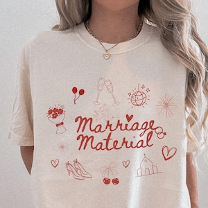 May include: A white t-shirt with the text "Marriage Material" in red lettering. The shirt is decorated with red illustrations of wedding-related items, including a bouquet, champagne flutes, a church, hearts, and balloons.