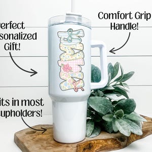 Puede incluir: Vaso blanco con asa y dise&ntilde;o floral "Mama". El vaso tiene tapa blanca y pajita. Tambi&eacute;n se ve el texto "Perfect Personalized Gift!" y "Fits in most cupholders!"