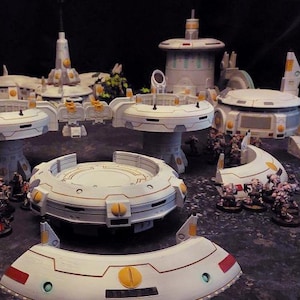 May include: A miniature sci-fi terrain set featuring white and gray structures with yellow and red accents. The set includes circular platforms, buildings, and curved elements, arranged on a dark surface, likely for tabletop gaming.