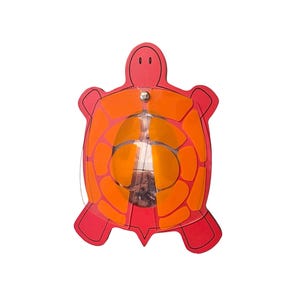 Può includere: Un acchiappasole a forma di tartaruga rossa e arancione. L'acchiappasole ha un design a strati con un centro trasparente e un fermaglio in metallo. Il guscio della tartaruga è arancione e il corpo è rosso.