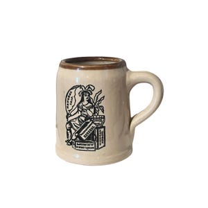 May include: A beige ceramic mug with a brown rim and handle. The mug features a black and white illustration of a woman and text that reads "Natural Leaf" and "Kavanah".