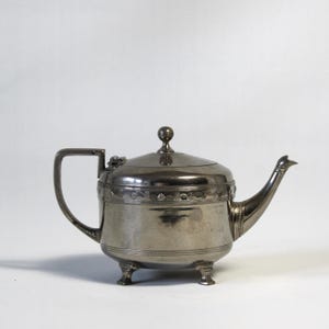 Op de afbeelding: Een theepot van gepolijst zilverkleurig metaal met een ronde behuizing, een gebogen tuit en een plat handvat. Het deksel is bekroond met een kleine bolvormige knop. De theepot rust op drie korte, stevige pootjes en heeft een decoratieve geometrische band.