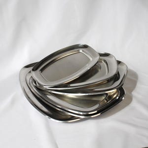 May include: A stack of stainless steel serving trays. The trays are oval-shaped with rounded corners and a polished, reflective surface. The trays are nested together, showcasing their varying sizes. The background is a plain white surface.