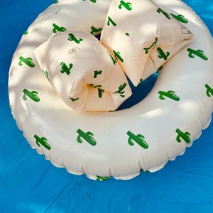 May include: A white inflatable pool ring with green cactus patterns, and two matching armbands. The items are on a blue surface, possibly a pool. The ring float is designed for water use.