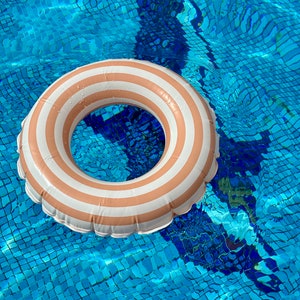 May include: A white and tan striped inflatable pool ring floating in a blue swimming pool. The water's surface reflects light, and the pool's tiled bottom is visible. The ring is round and has a classic design.