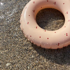 May include: A pale pink inflatable ring with a cherry pattern, partially in water on a pebbled beach. The cherries are small and red, dotted across the surface. The ring is a summer accessory for the beach or pool.
