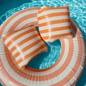 May include: An inflatable pool ring and two matching pillows floating in a pool. The ring and pillows feature orange and white stripes. The water is a clear, bright blue.