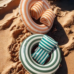 May include: Two inflatable pool floats and two matching pillows rest on sand. One float is orange and white striped, the other is green and white striped. The pillows have matching stripes. Ideal for a day at the beach or pool.