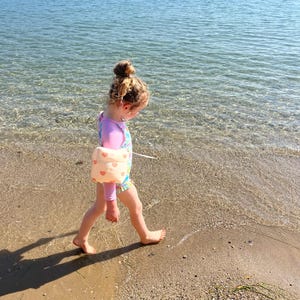 May include: A child in a pink and patterned swimsuit and arm floaties walks on a sandy beach. The child has their hair in a bun. The water is clear and the sky is blue.