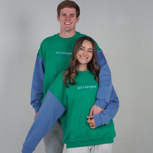 May include: Two people wearing matching green and blue color block sweatshirts. The sweatshirts have the text "let's kozyup" in light blue script. The background is a solid light gray.