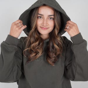 May include: Dark olive green hoodie with a textured hood and the word "KOZY" embroidered on the front. The hoodie has long sleeves and a relaxed fit. The model is wearing the hood.