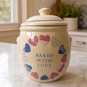 May include: A cream-colored ceramic cookie jar with a matching lid, featuring a circular pattern of blue and pink heart-shaped patches. The text "BAKED WITH LOVE" is printed in blue letters in the center of the heart arrangement on the front.