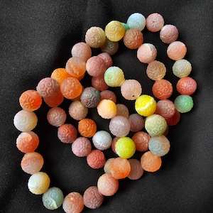 May include: A selection of colourful, carved gemstone beads in shades of orange, pink, yellow, green, and blue. The beads are round with a detailed, swirling pattern. The beads are displayed on a black background.
