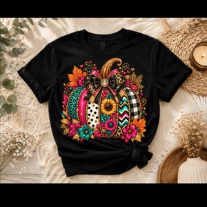 Könnte beinhalten: Ein schwarzes T-Shirt mit einer zentralen Grafik eines Kürbisses, der mit verschiedenen Mustern wie Leopardenmuster, Punkten, Sonnenblumen und Karos verziert ist. Der Kürbis ist mit einer großen schwarzen Schleife und Herbstblättern geschmückt.