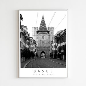 May include: Black and white framed print of Basel, Switzerland. The image features a street scene with a large stone gate and buildings on either side. The word "BASEL" and coordinates are printed at the bottom.