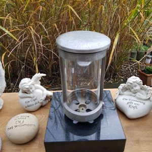 Könnte beinhalten: Eine silberfarbene Gedenklaterne mit Glaseinfassung, auf einem schwarzen Granitsockel. Die Laterne ist von dekorativen Engelsfiguren und Gedenksteinen mit deutschem Text umgeben, wodurch eine ruhige und gedenkende Darstellung entsteht.