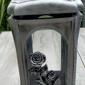 May include: A silver metal lantern with a glass window featuring a rose design. The lantern has a textured surface and a hinged lid.